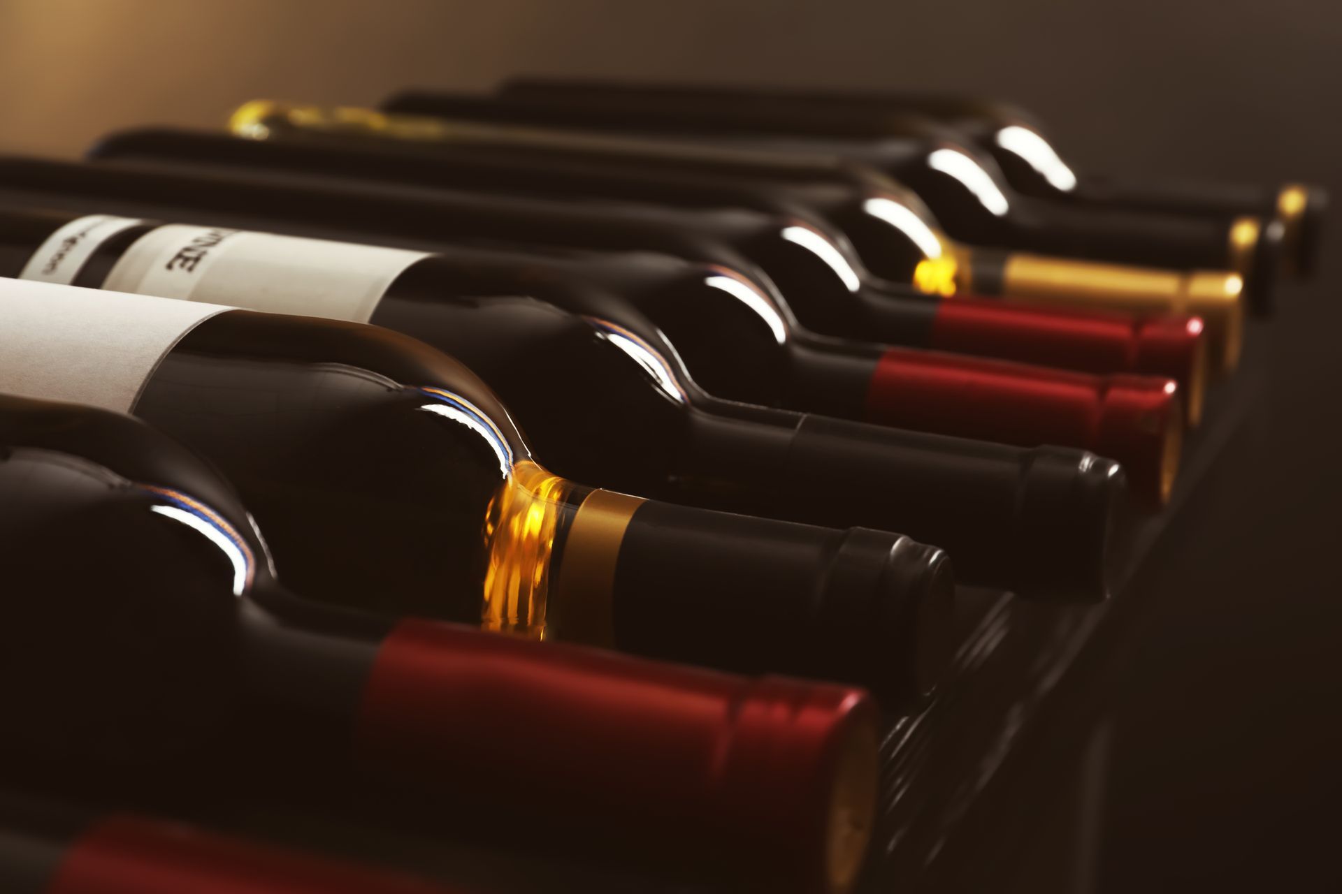 Des rangées de bouteilles de vin couchées sur le côté dans une cave faiblement éclairée, dont on distingue les étiquettes et les verres colorés.