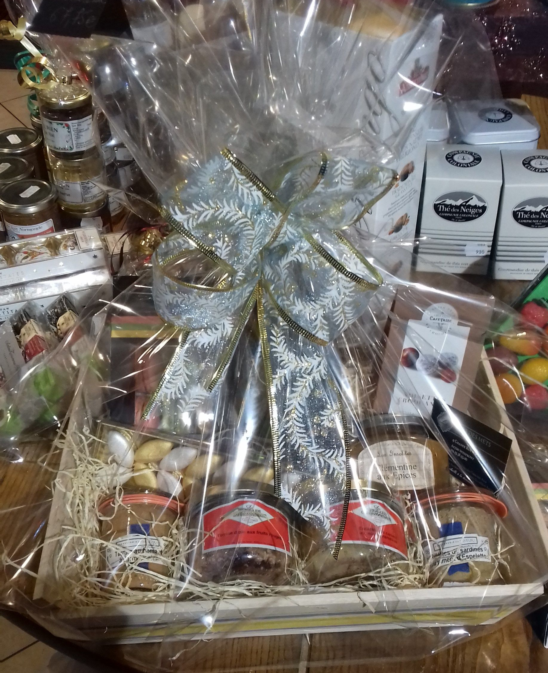 Panier cadeau contenant des bocaux assortis et des produits emballés, le tout enveloppé dans du papier transparent avec un ruban.