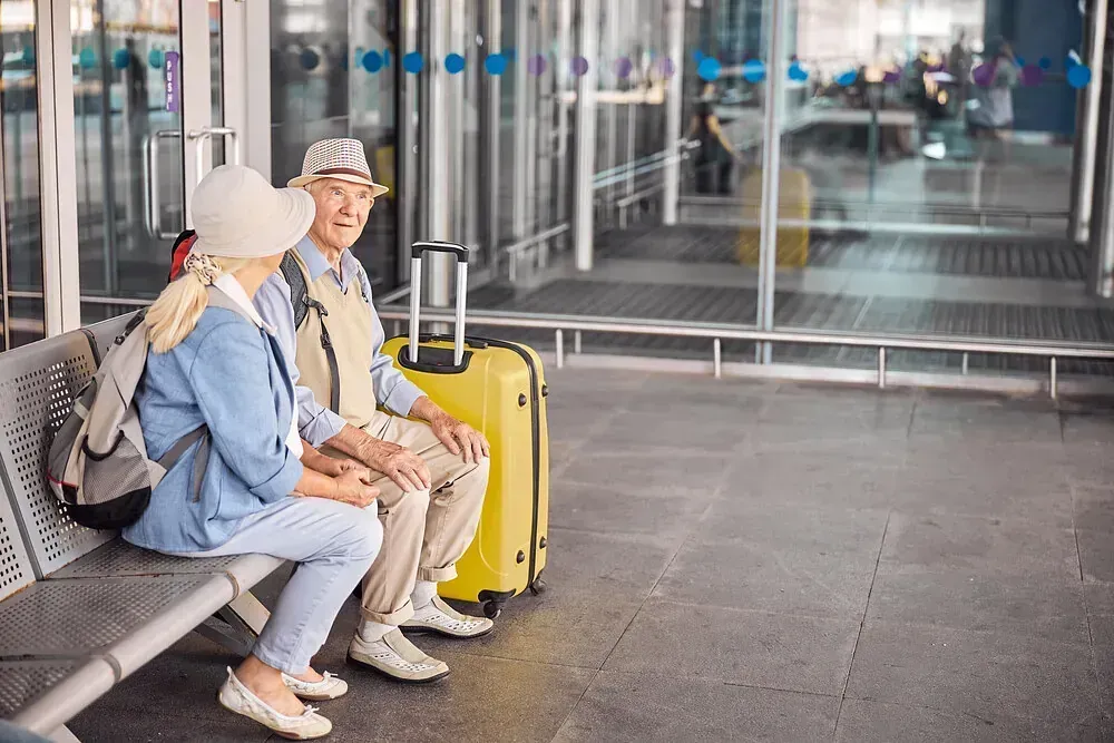 Pareja de ancianos en un aeropuerto, sentados en un banco junto a un equipaje amarillo, con sombreros, hablando.