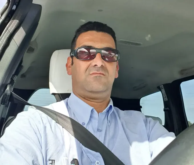 Un hombre que lleva gafas de sol y una camisa azul en un automóvil, abrochado el cinturón de seguridad. Tiene una expresión neutra y el entorno parece ser de día.