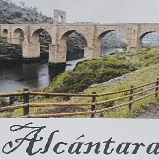 Puente romano en Alcántara, España, con arcos sobre un río. En primer plano, una valla de madera y una ladera.