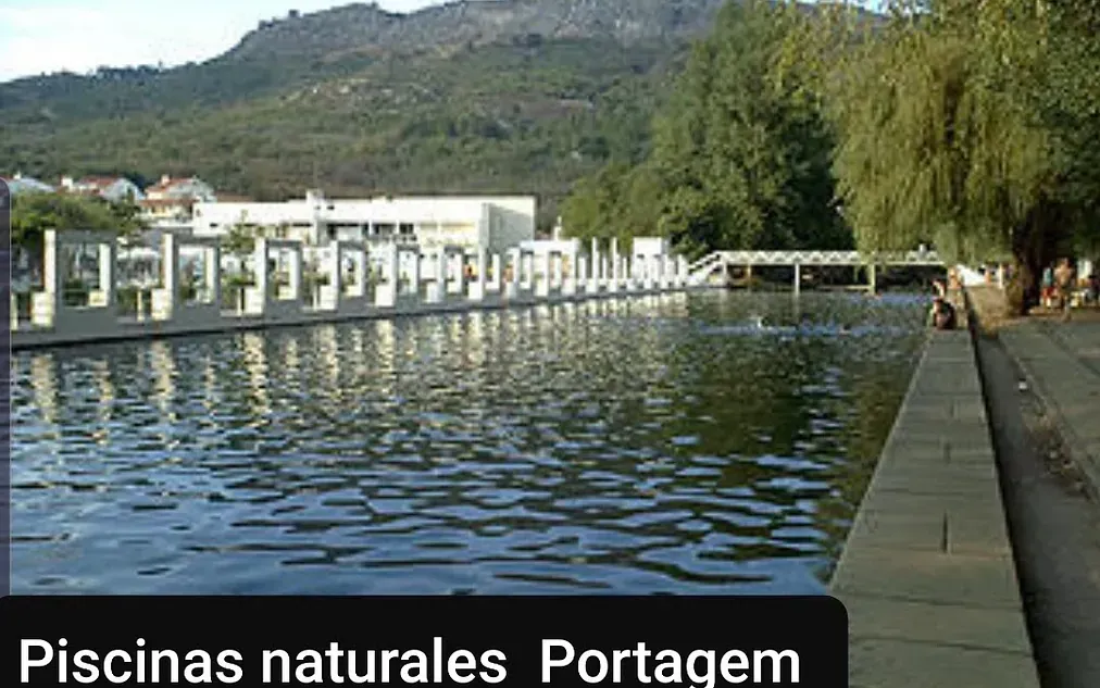 Piscinas naturales en Portagem, Portugal. Una larga masa de agua rectangular está bordeada por una pasarela de piedra y estructuras blancas. Montaña al fondo.