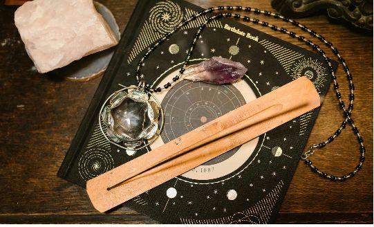 Varita de incienso sobre una tabla negra con motivos celestiales, con cristal, amatista y cadena sobre una mesa de madera.