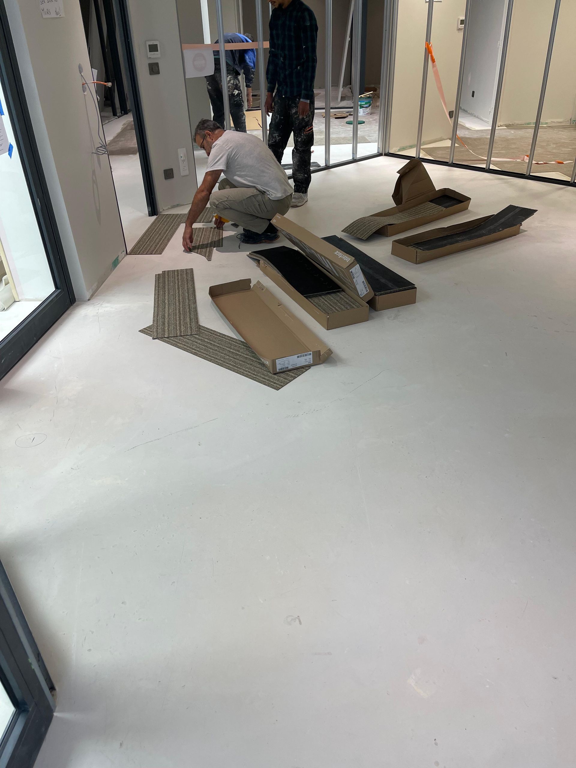 Un homme à genoux pose du carrelage dans une pièce en construction, des cartons de carreaux se trouvant à proximité.