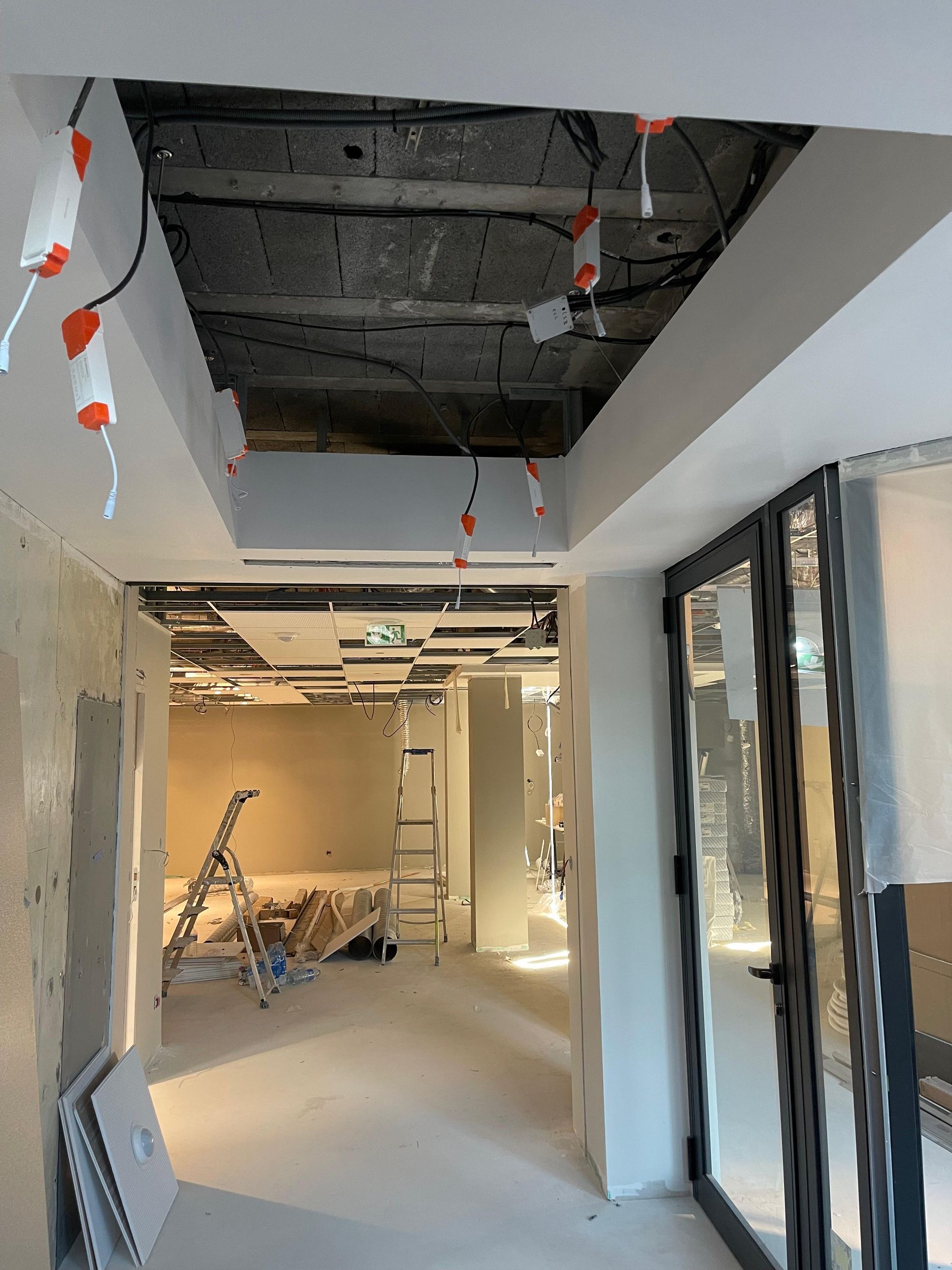 Couloir intérieur en travaux ; câbles apparents au plafond, éclairage encastré, murs non finis et portes vitrées.