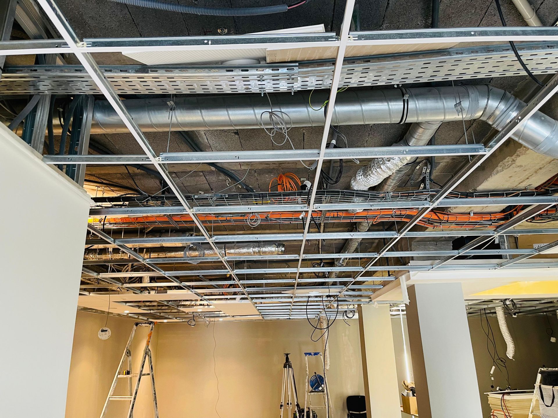 Chantier de construction avec faux plafond, conduits et câbles apparents. On aperçoit des murs blancs et une échelle.