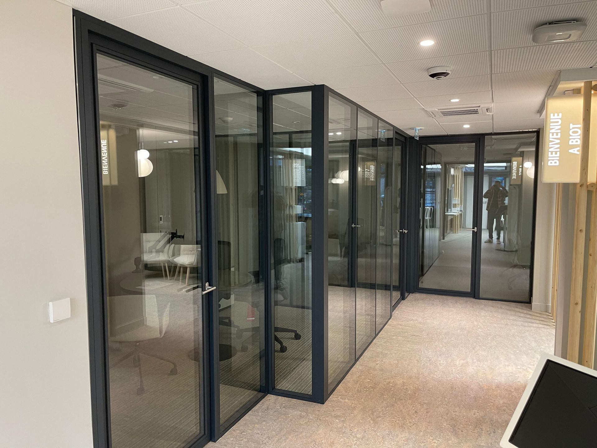 Couloir intérieur avec bureaux aux parois vitrées. Encadrements et portes gris, moquette beige et plafond blanc.