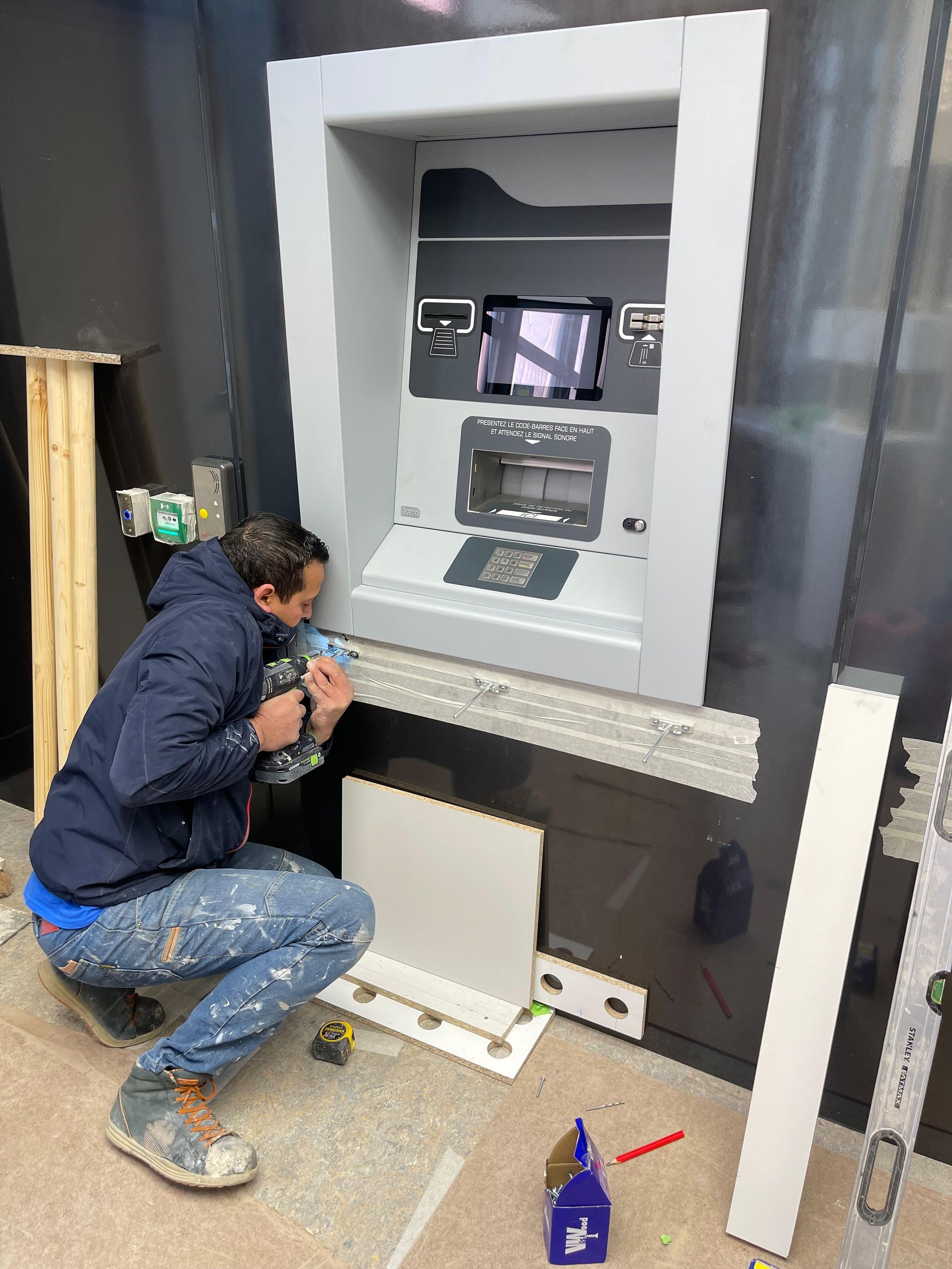 Un homme installe un distributeur automatique de billets sur un mur sombre. Il est agenouillé et travaille avec des outils.