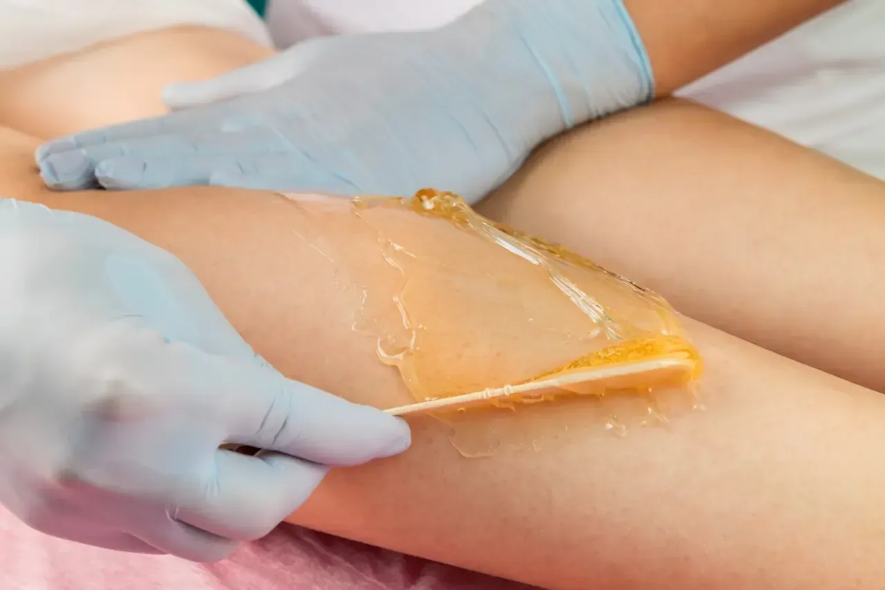 Pierna de una persona siendo depilada con cera. Una mano enguantada aplica la cera con una espátula de madera.