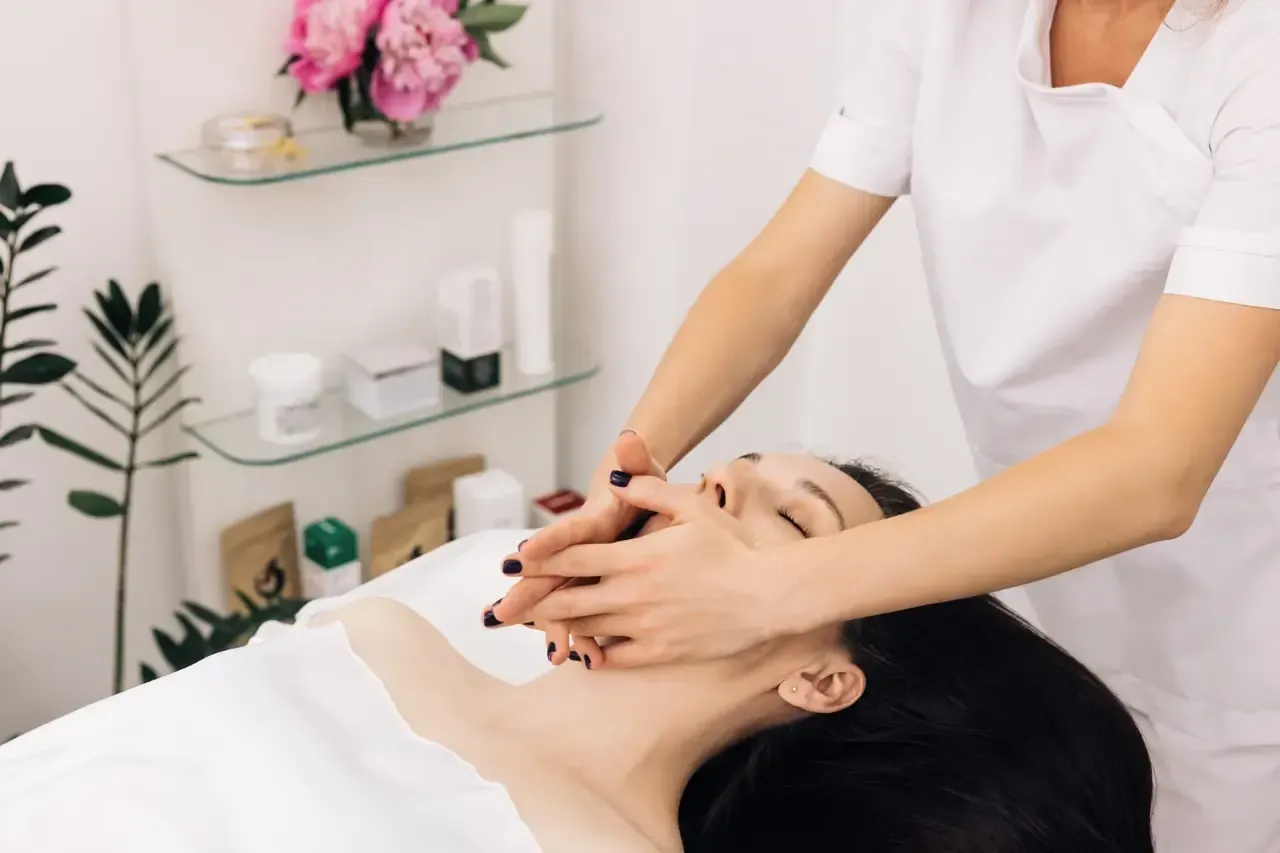 Mujer recibiendo un masaje facial en un spa. Manos sobre su rostro, en una habitación luminosa.