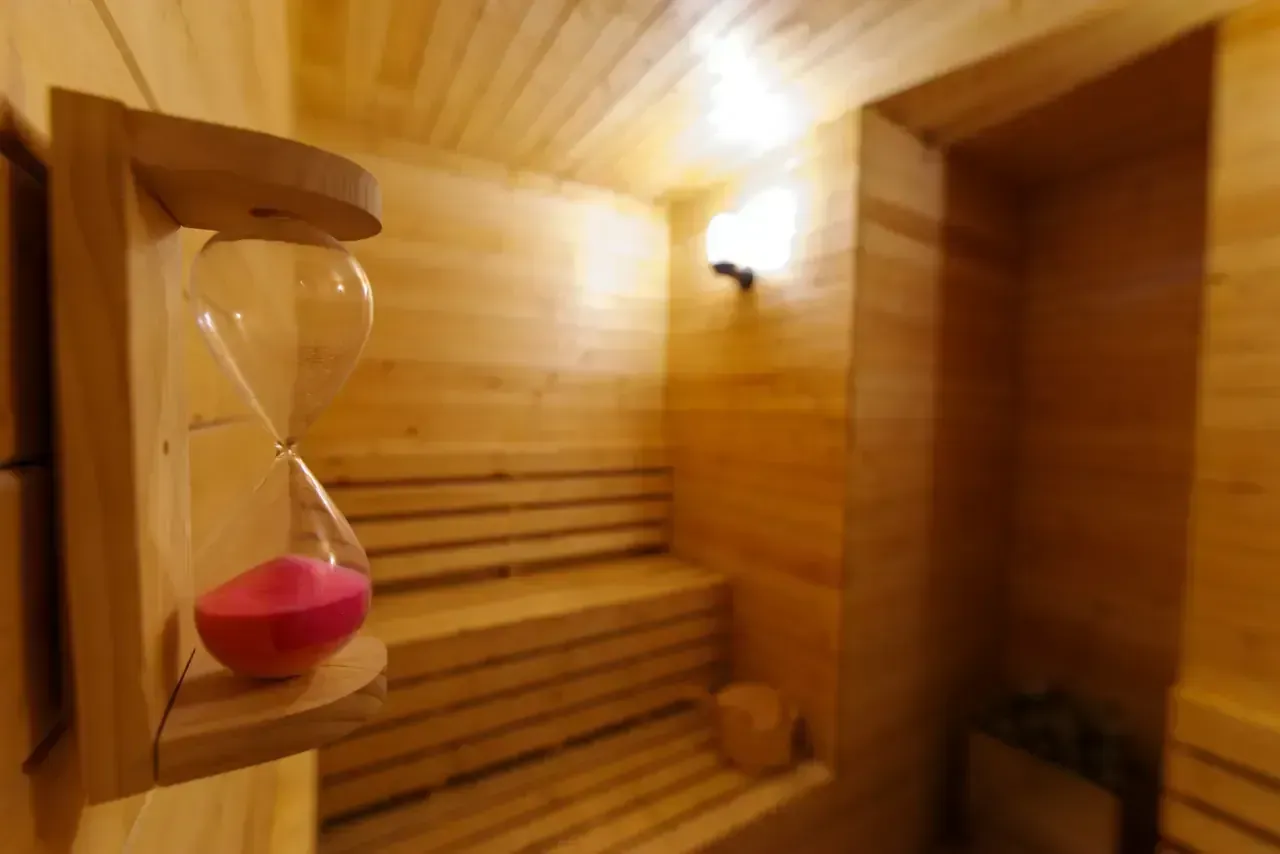 Interior de sauna de madera con reloj de arena y bancos. Paredes y grifería de madera clara.