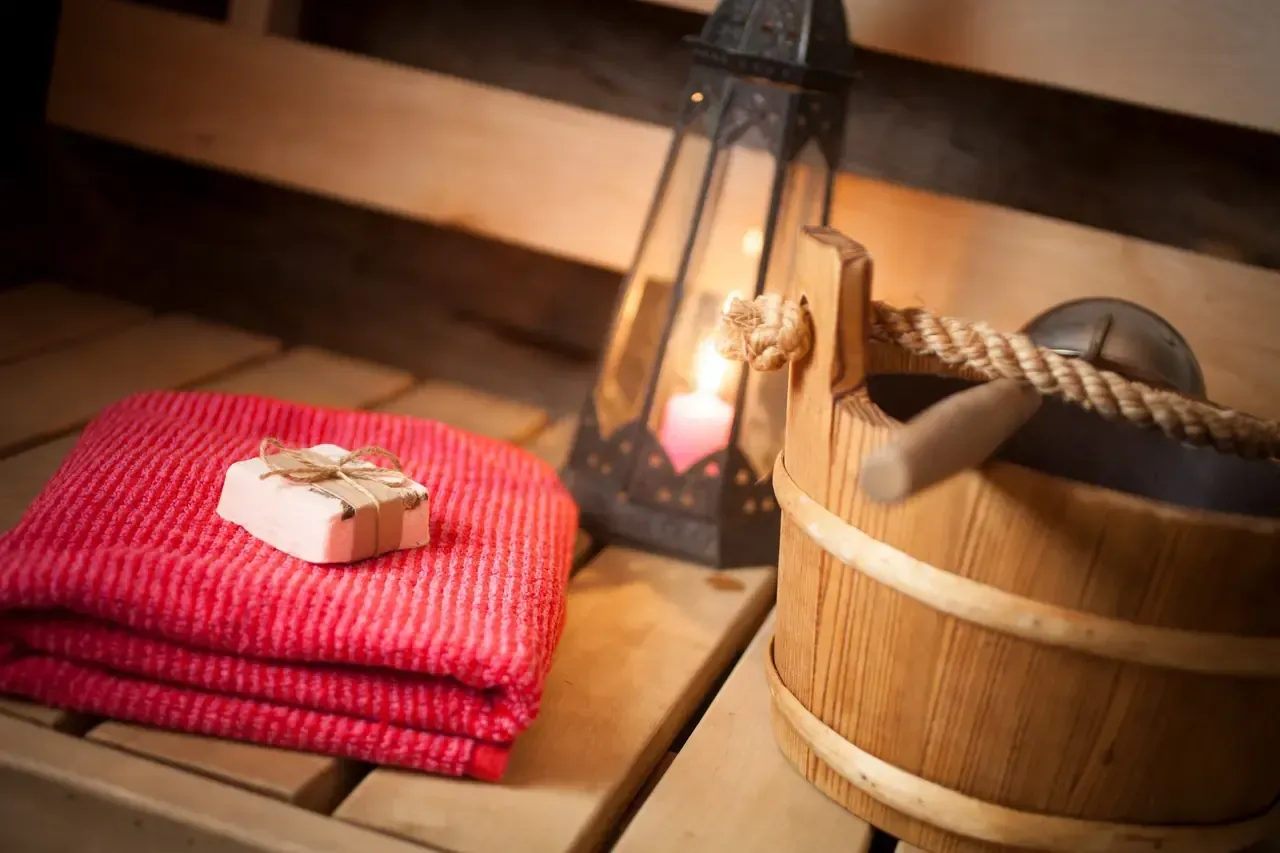 Toalla roja con jabón, cubo de madera, farol con vela sobre un banco de sauna.