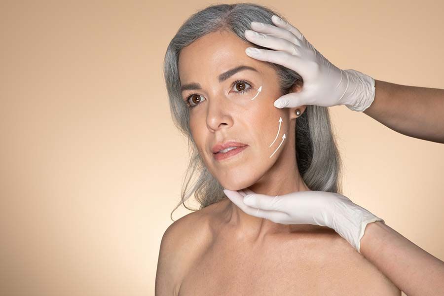 Rostro de mujer con marcas quirúrgicas; manos enguantadas examinan su rostro, fondo beige.