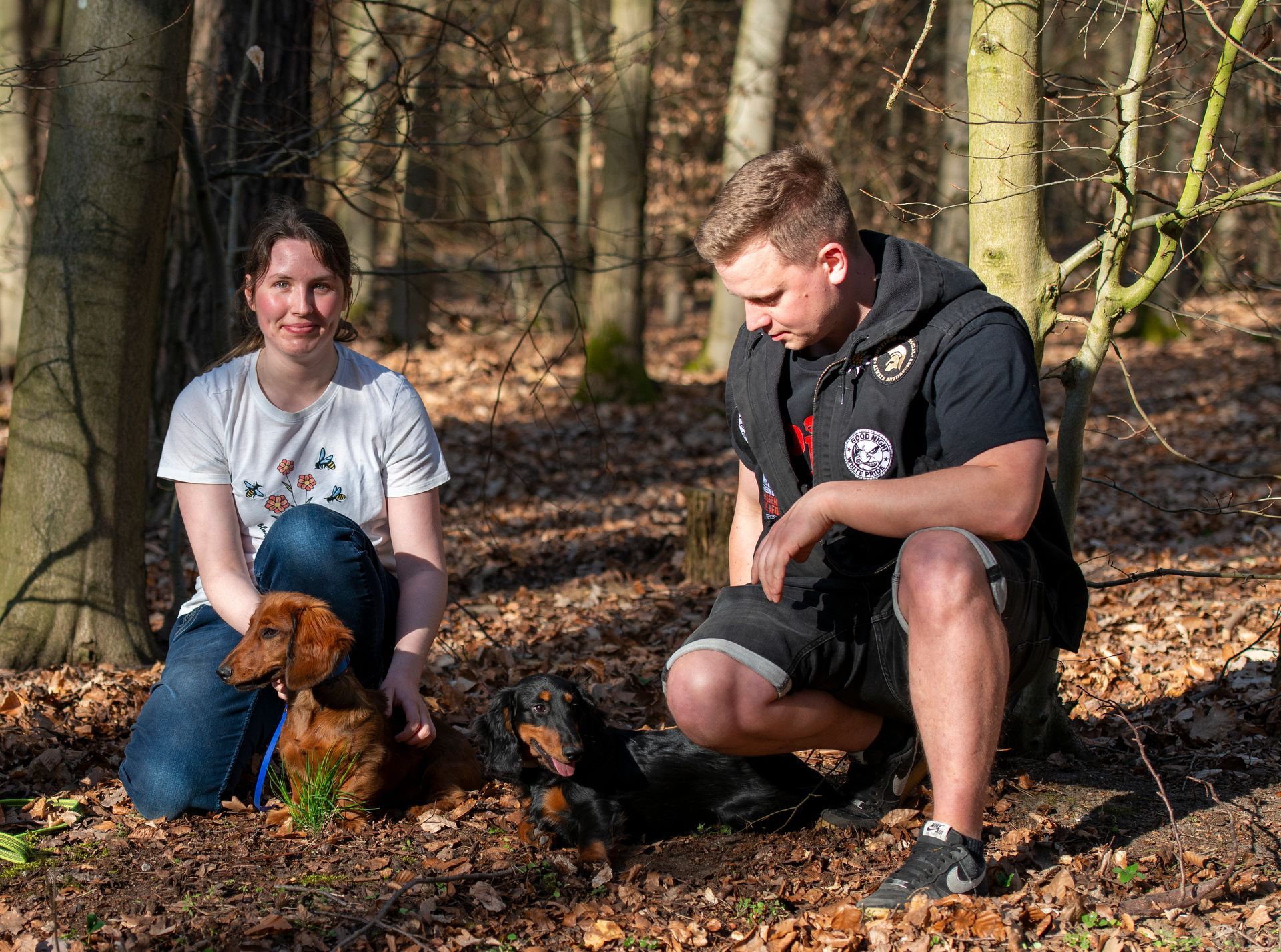 Ein Mann und eine Frau sitzen mit ihren Dackeln im Wald.
