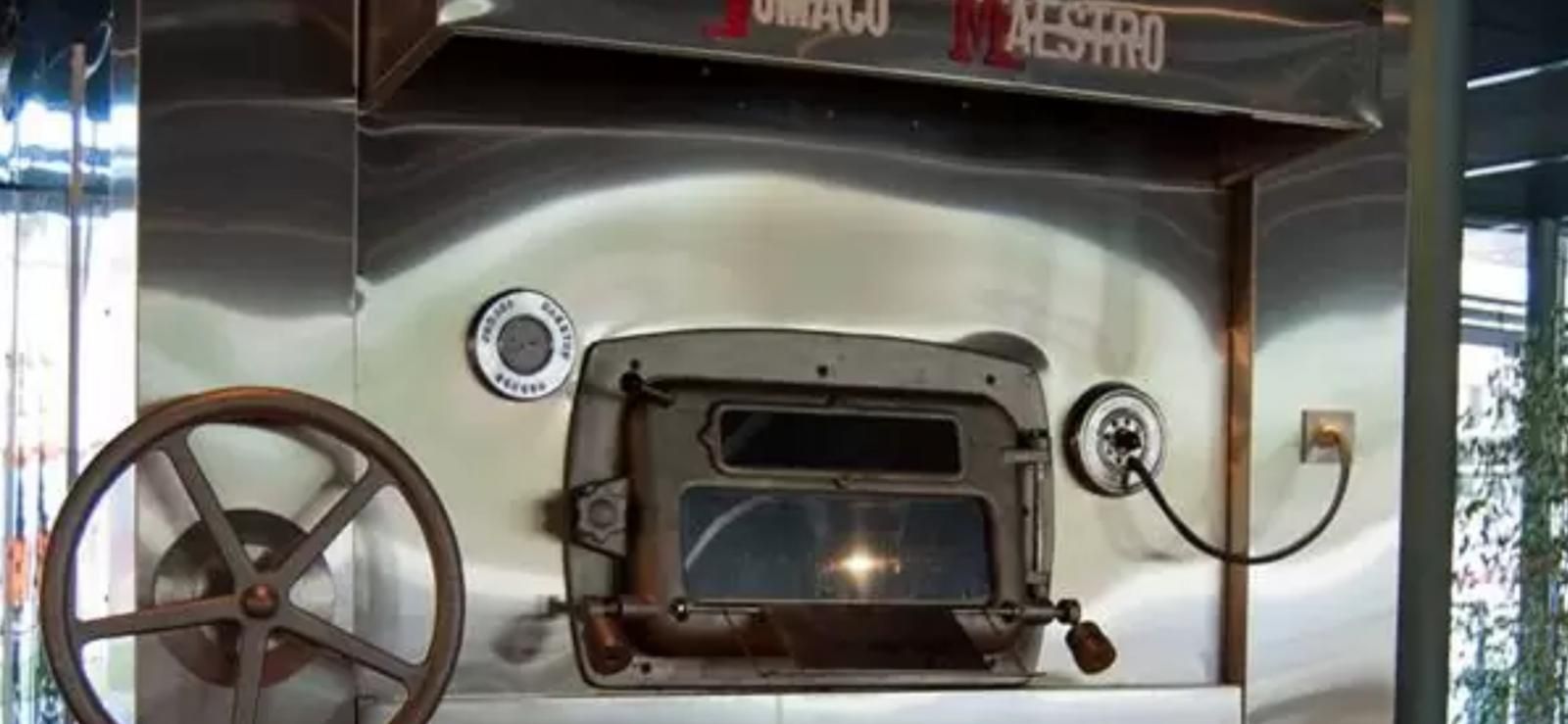 Horno de pizza de acero inoxidable con puerta oscura y abierta. Rueda metálica a la izquierda.