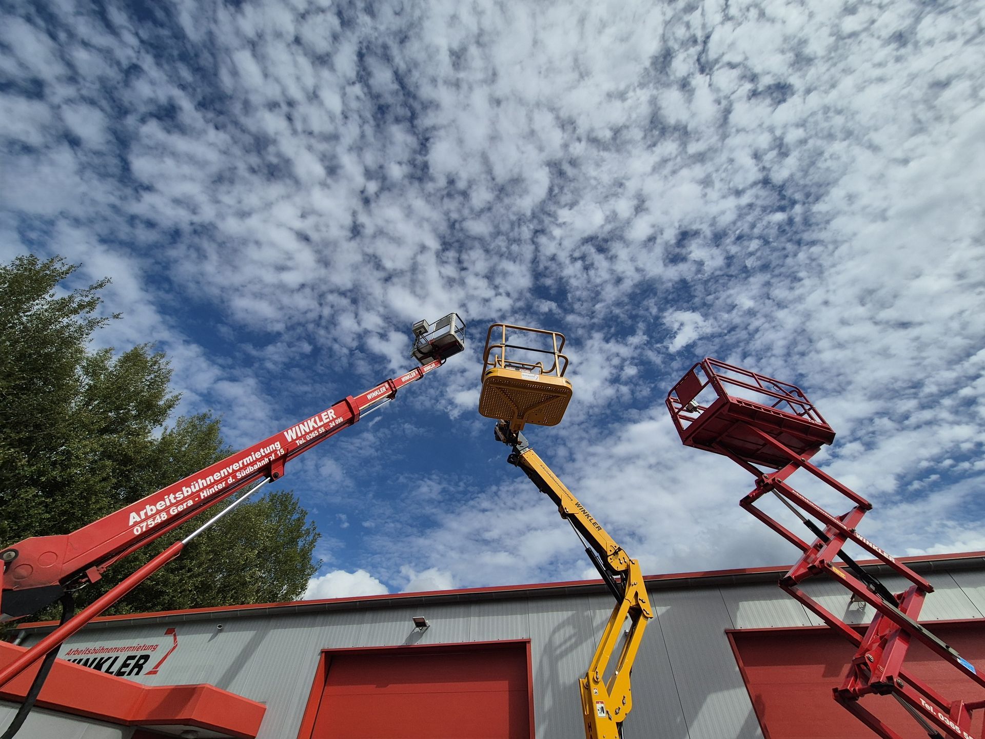 Zwei Hubarbeitsbühnen, eine orange und eine blau, vor einem strahlend blauen Himmel mit Wolken.