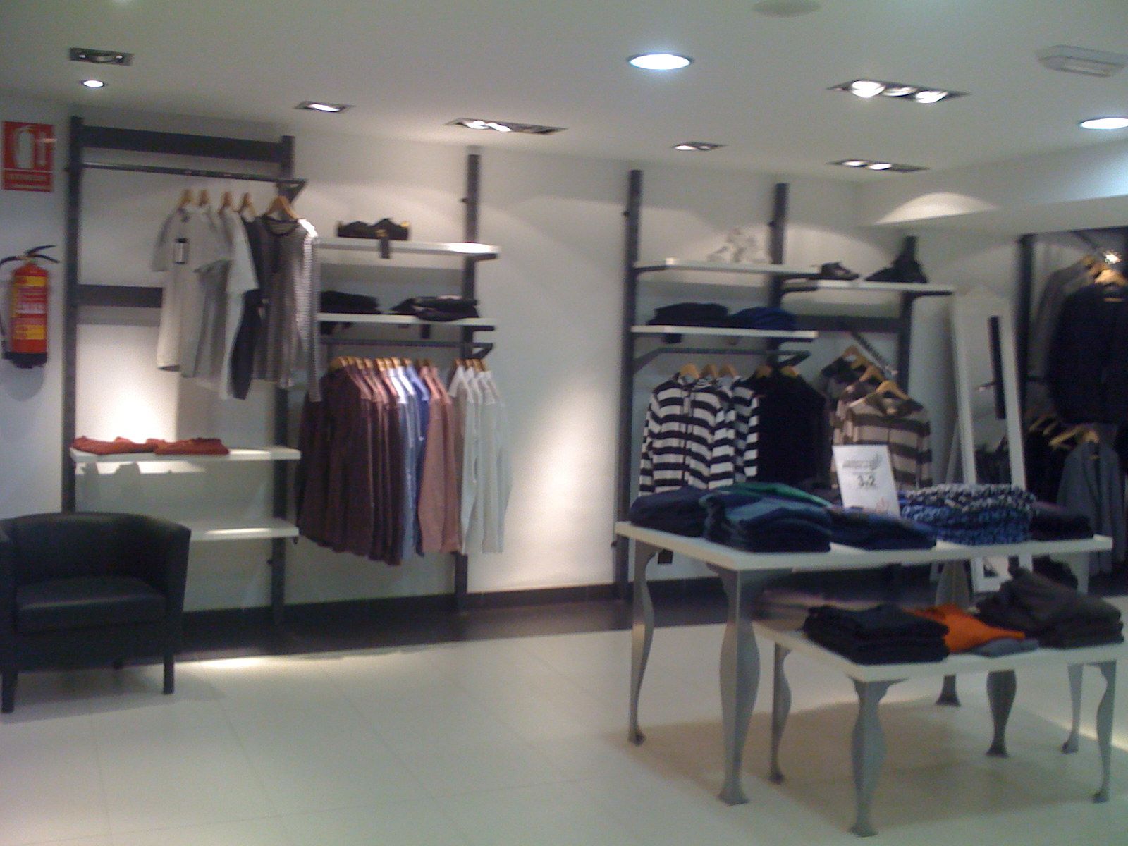 Interior de una tienda de ropa con percheros llenos de camisas y suéteres doblados bajo luces brillantes del techo.