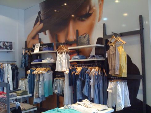 Exhibición de una tienda de ropa con jeans colgados, prendas blancas y un gran póster de moda sobre los estantes.