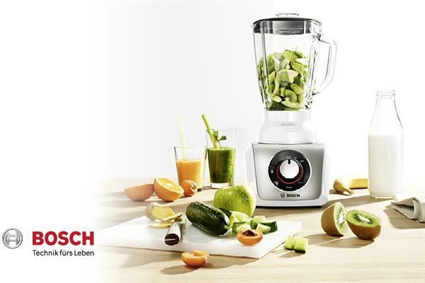 Auf einem Holztisch steht ein Mixer, umgeben von Obst und Gemüse.