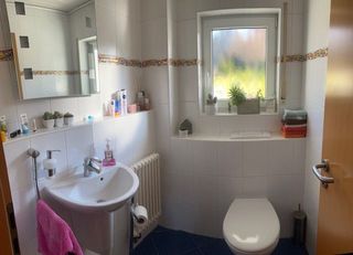 Kleines Badezimmer mit Waschbecken, Toilette, Fenster, Spiegel und Toilettenartikeln auf der Ablage.