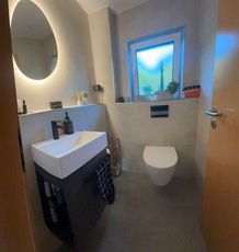 Modernes Badezimmer mit wandmontiertem Waschbecken, rundem Spiegel, Toilette und kleinem Fenster mit Blick auf den blauen Himmel.