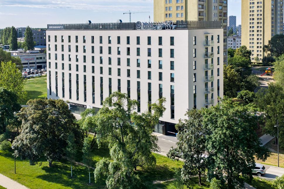 Nowoczesny biały budynek biurowy otoczony drzewami, w tle wysokie wieżowce mieszkalne.