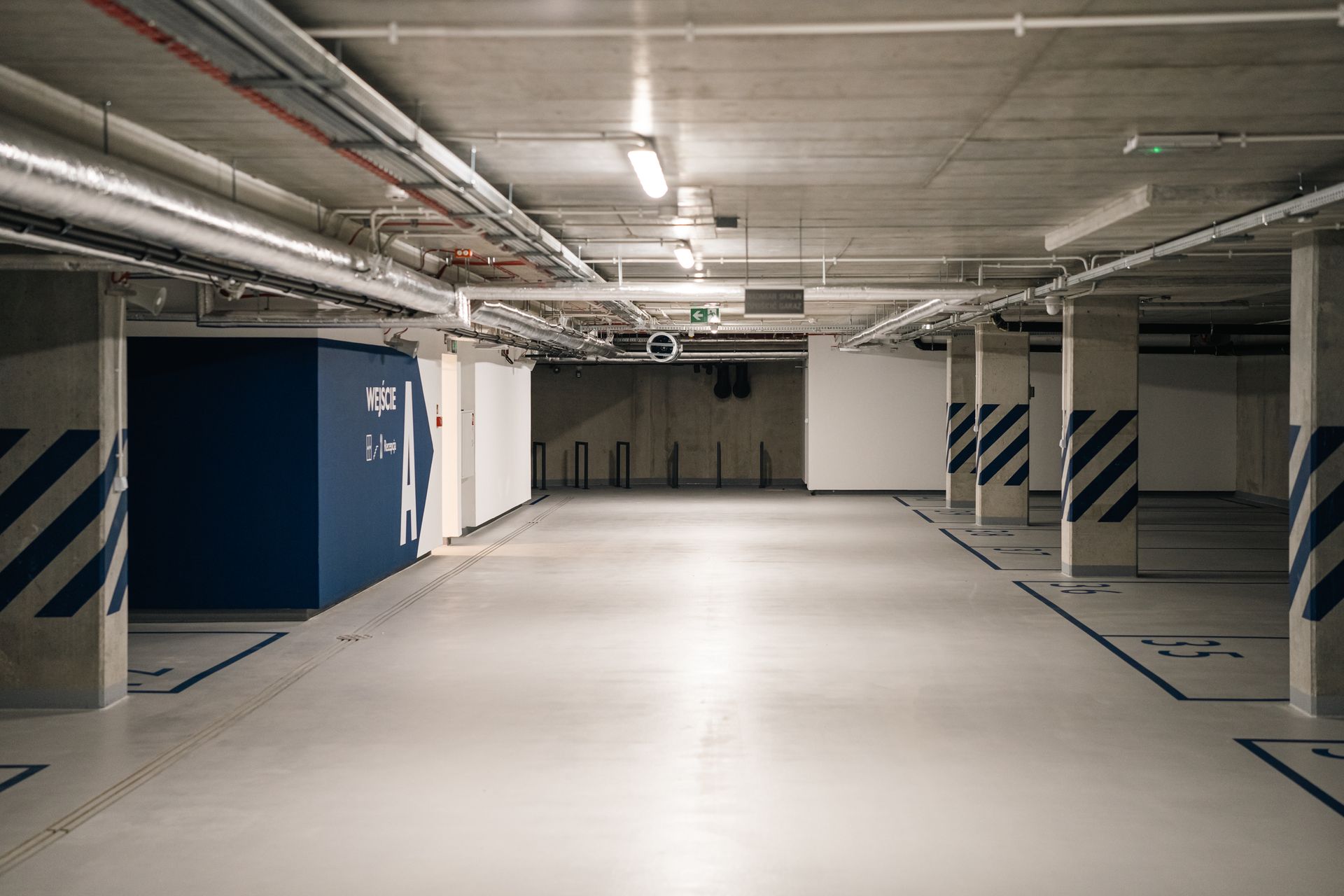 Pusty podziemny parking z białą podłogą, betonowymi filarami i niebieskimi szafkami gospodarczymi