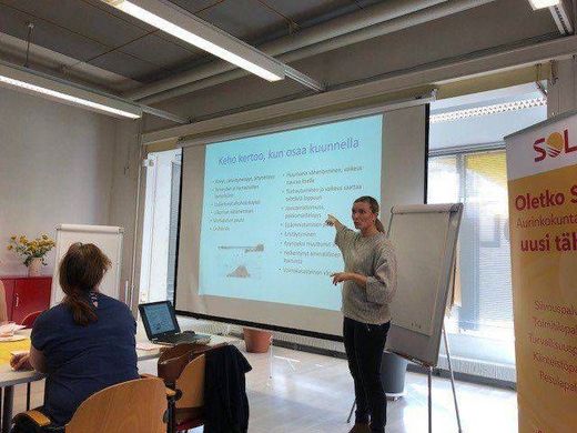 Esittäjä puhuu projisoidun dian vieressä kirkkaassa luokkahuoneessa; osallistuja istuu kannettavan tietokoneen ja fläppitaulun ääressä lähellä