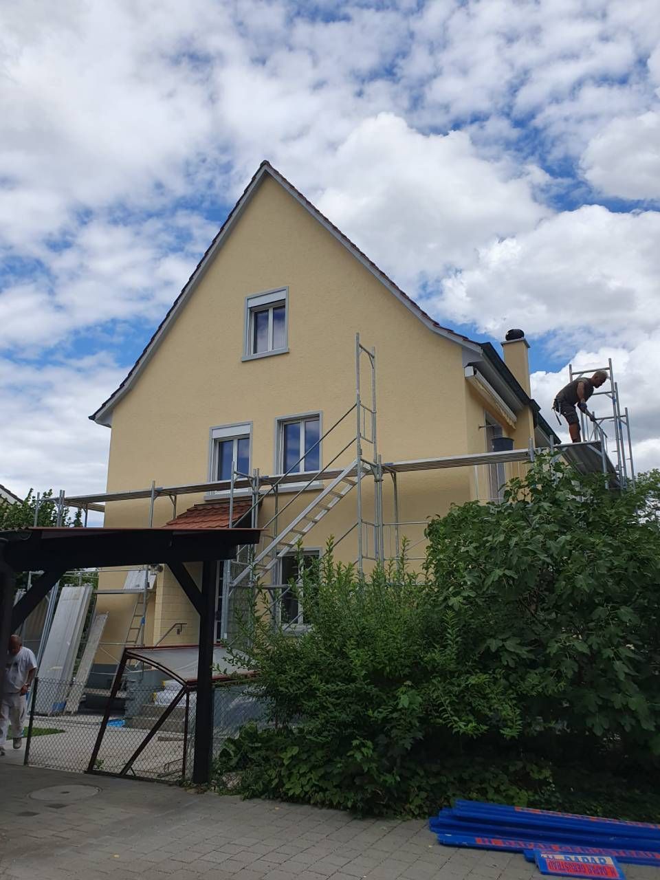 Die Hausfassade wird renoviert, ein Gerüst steht. Gelbe Verkleidung, eine Person auf dem Dach.