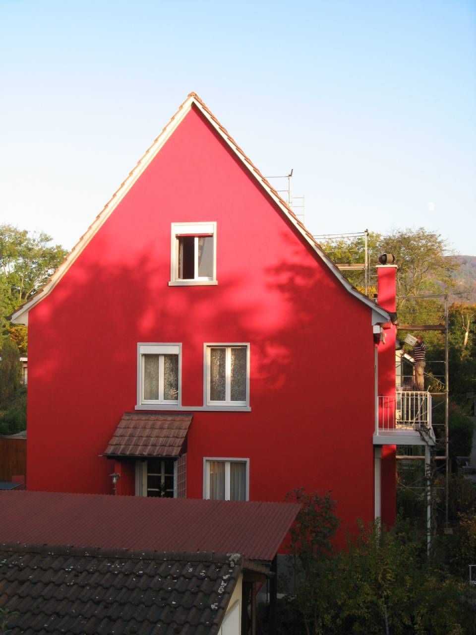 Ein leuchtend rotes Haus mit weiss umrahmten Fenstern vor blauem Himmel und einem roten Dach im Vordergrund.