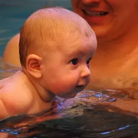 Ein Baby treibt mit dem Gesicht über Wasser in einem Pool; ein Erwachsener ist in der Nähe.