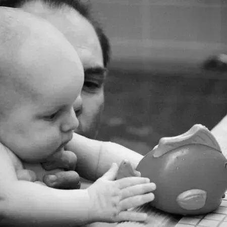 Auf einem Schwarz-Weiß-Foto greift ein Baby nach einem Spielzeug, während ein Erwachsener es fest an sich drückt.