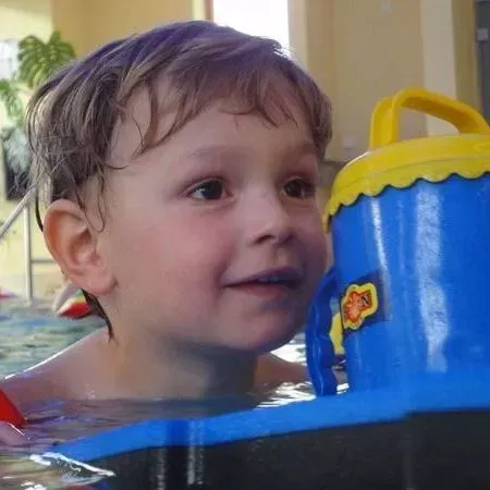 Kind im Pool, das einen blauen Spielzeugeimer mit gelbem Henkel hält, blickt nach oben und lächelt.