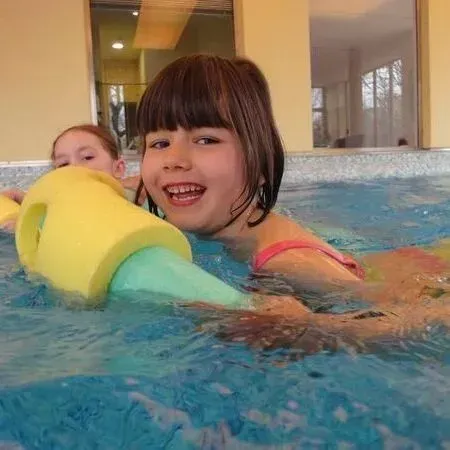 Ein lächelndes Kind hält in einem Schwimmbecken eine gelbe Schwimmhilfe; im Hintergrund ist ein weiteres Kind zu sehen.