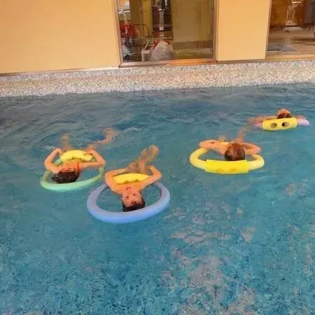 Menschen treiben auf bunten Schwimmreifen in einem Pool; im Hintergrund sind ein Gebäude und eine Tür zu sehen.