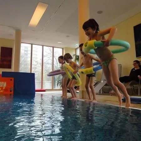 Kinder in Schwimmringen beugen sich über ein Hallenbad und greifen nach dem Wasser