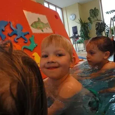 Kinder in einem bunten Hallenbad, eines lächelt in die Kamera, während an der Wand Wasserspielzeug hängt.