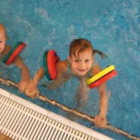 Zwei Kinder in einem Schwimmbecken, die bunte Schwimmflügel tragen, sich am Beckenrand festhalten und lächeln.