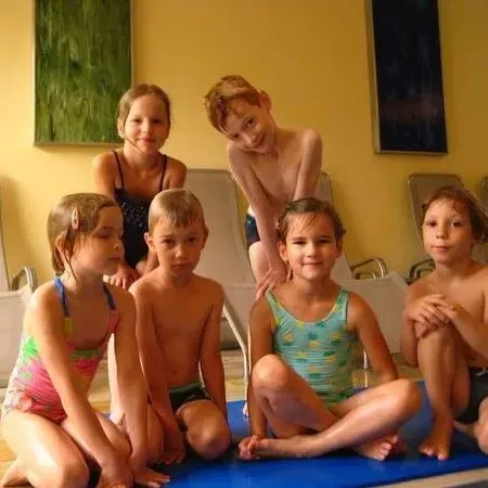 Fünf Kinder sitzen auf blauen Matten in einem Raum, dahinter stehen zwei Frauen, an der Wand hängen abstrakte Gemälde.