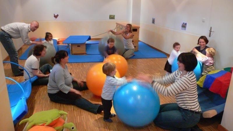 Menschen spielen in einem Therapieraum mit großen blauen und orangen Gymnastikbällen auf einem gepolsterten Boden.