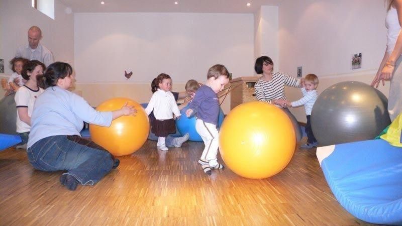 Kinder und Erwachsene spielen in einer Turnhalle mit großen orangefarbenen, grauen und blauen Gymnastikbällen.