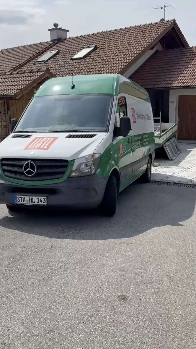 Ein weiß-grüner Mercedes-Transporter parkt vor einem Haus mit Ziegeldach in einer sonnigen Straße.