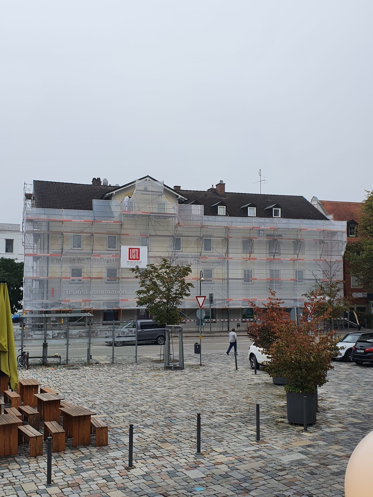 Ein eingerüstetes Gebäude wird in einem verschneiten Innenhof renoviert; Bänke und einige Fußgänger sind anwesend.