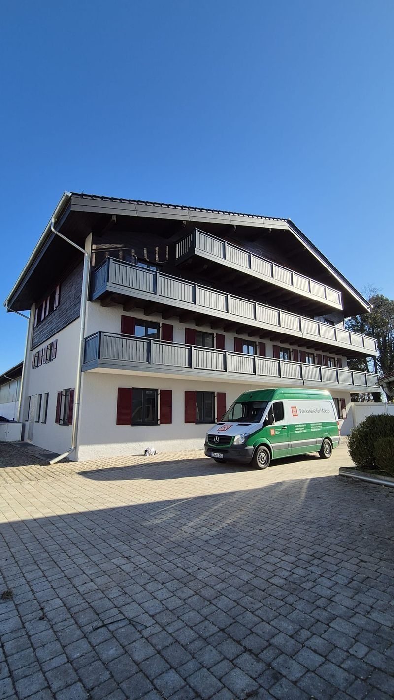 Ein Gebäude im Chalet-Stil mit Balkonen und Fensterläden, davor ein grüner Lieferwagen, auf einem sonnigen, gepflasterten Grundstück.
