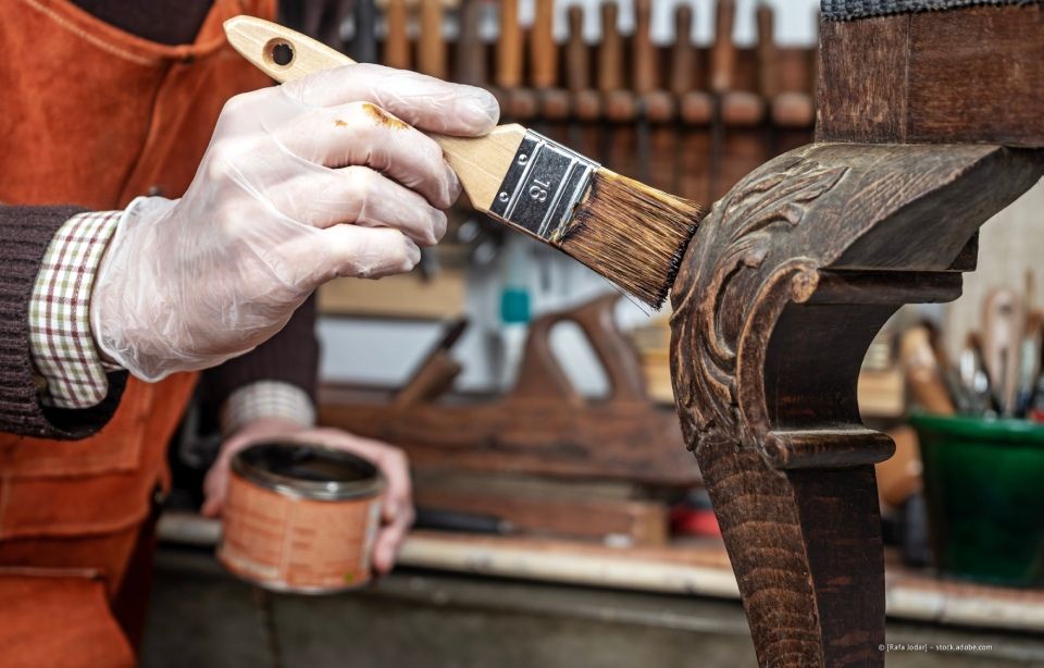 Eine behandschuhte Hand bemalt in einer Werkstatt das geschnitzte Bein eines Holzstuhls.