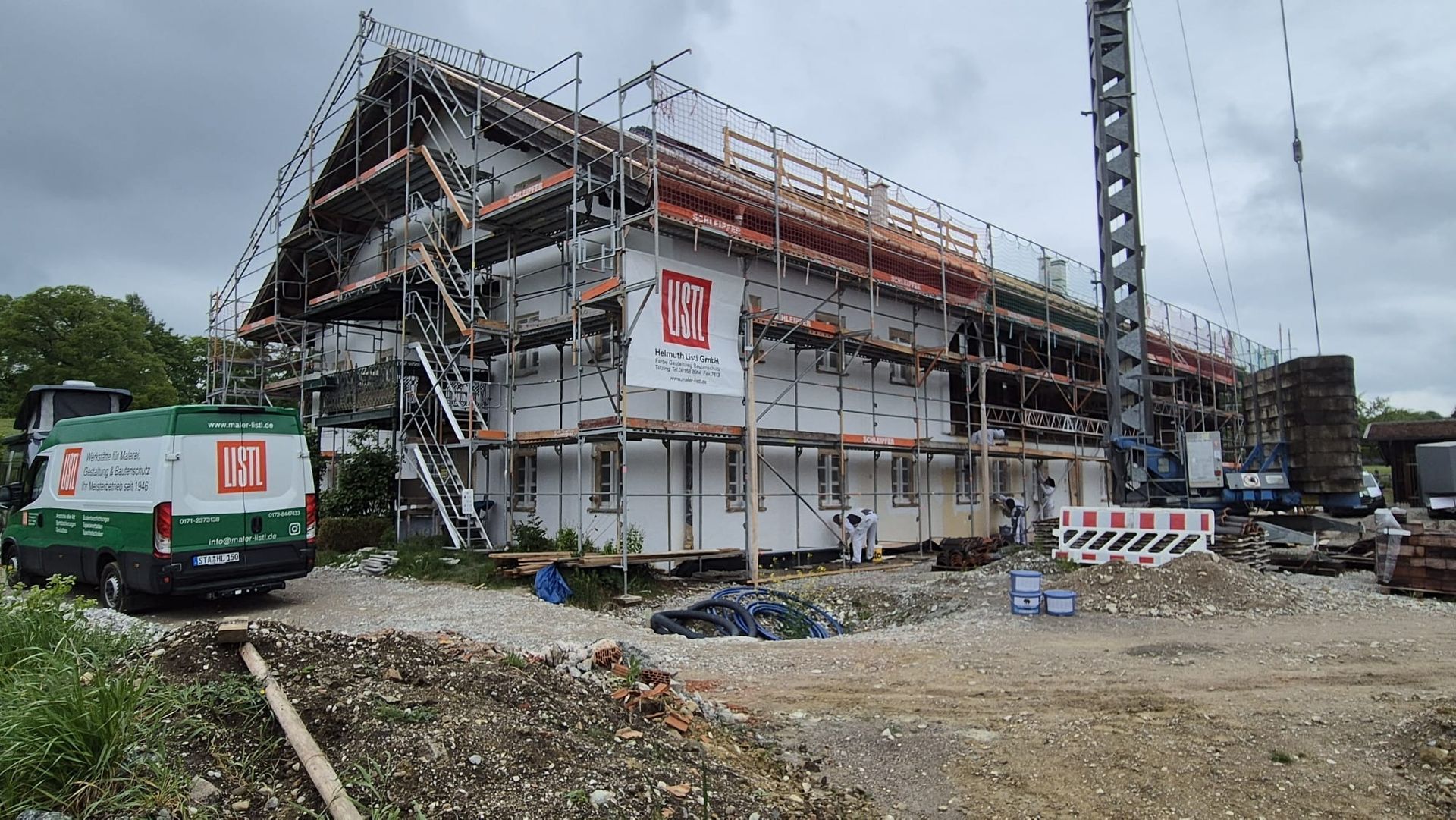 Ein mit einem Gerüst versehenes, im Bau befindliches Haus neben einer matschigen Baustelle, daneben ein Lieferwagen und Absperrungen.