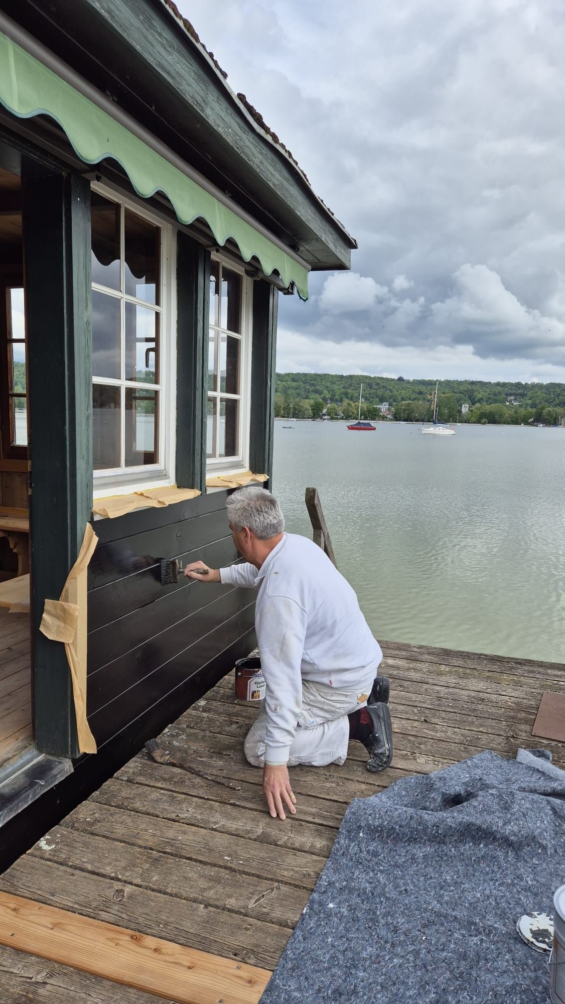 Ein Mann in weißer Kleidung greift in einen schwarzen Außenheizstrahler neben einer Hütte am See.