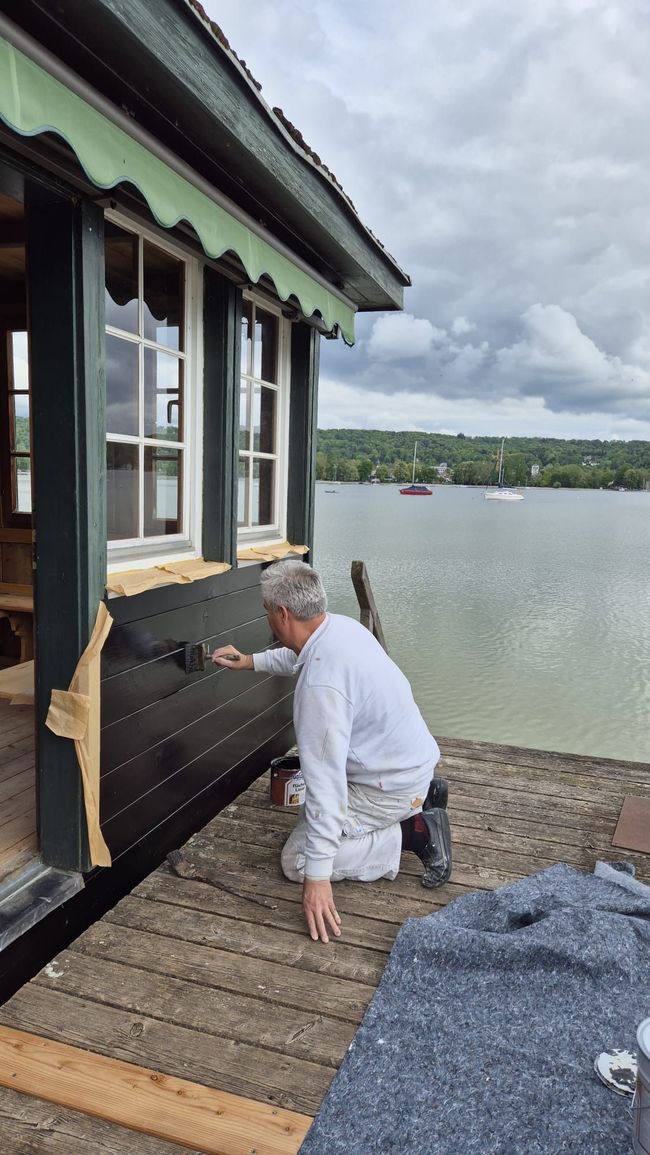 Ein Mann in weißer Kleidung greift in einen schwarzen Außenheizstrahler neben einer Hütte am See.