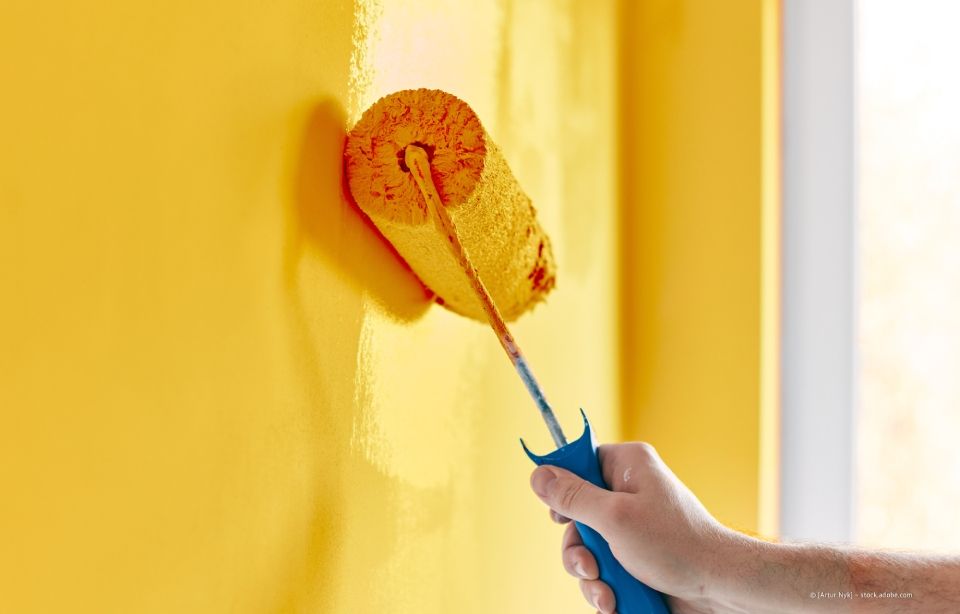 Eine gelbe Wand mit einer Farbrolle mit blauem Griff von Hand streichen.