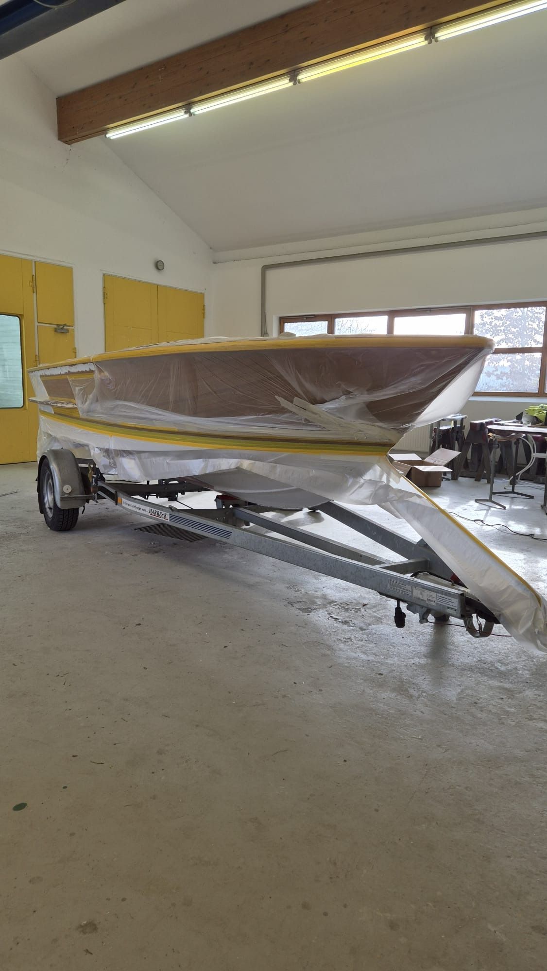 Ein eingewickeltes Boot auf einem Anhänger in einer Garage, mit gelben Schränken und Neonröhren an der Decke.